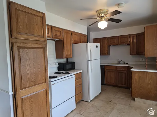 Kitchen featuring wood cabinetry, white appliances, stone-finish tile flooring, a white tile backsplash, and a ceiling-mounted light fixture with an integrated fan