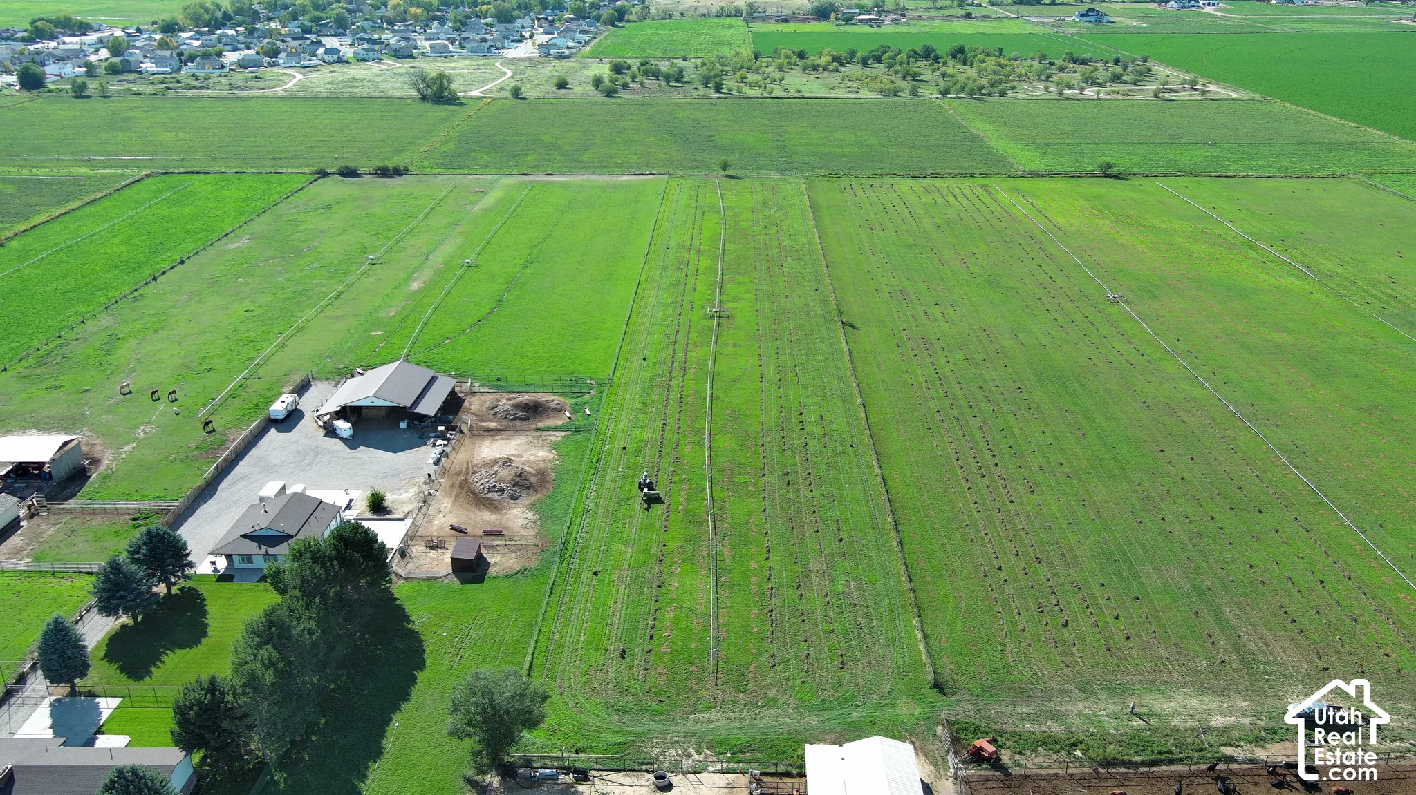 Expansive green acreage with planted rows, featuring a structure with a dark roof and a separate building with a light-colored roof, surrounded by paved and unpaved areas
