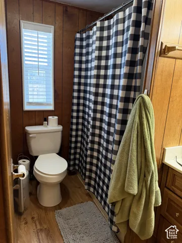Bathroom featuring wood-finish paneling, wood-finish flooring, a white toilet, and a window with blinds