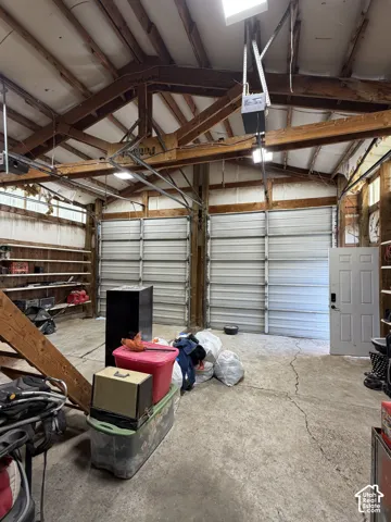 Exposed wood beam ceiling structure with skylight, two large roll-up doors, concrete flooring, and built-in shelving