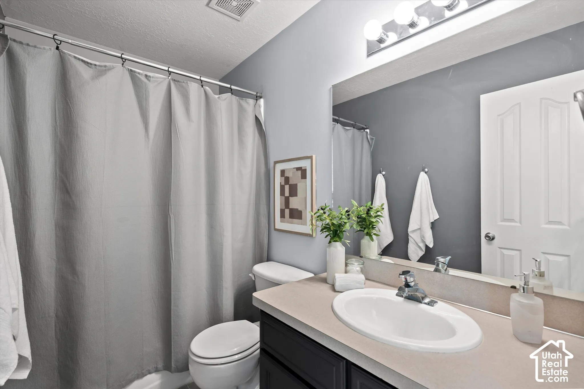 Bathroom featuring a single vanity with an oval sink, a light-toned countertop, a large wall-mounted mirror, overhead vanity lighting, and a white toilet
