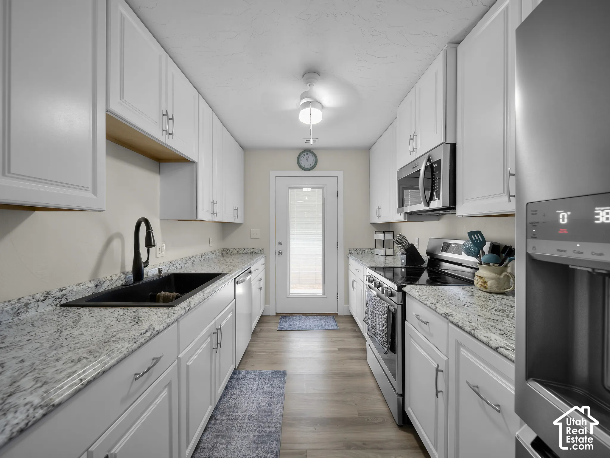Galley kitchen featuring white cabinetry, light-toned countertops, stainless steel appliances, a black basin sink with a matte black faucet, and wood-finish flooring