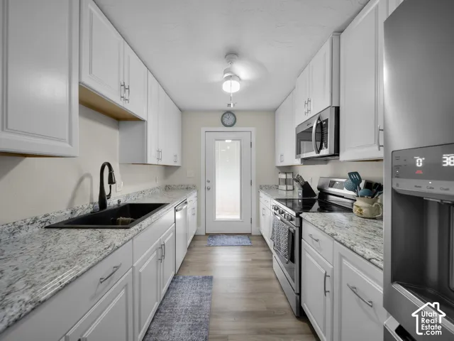 Galley kitchen featuring white cabinetry, light-toned countertops, stainless steel appliances, a black basin sink with a matte black faucet, and wood-finish flooring