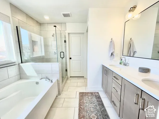 White tile flooring, a built-in bathtub, a glass-enclosed shower, a double vanity with light wood-finish cabinetry, and a large wall-mounted mirror