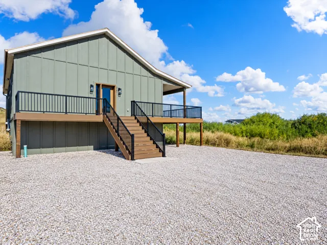 Board and batten siding, elevated deck with dark railings, wood-finish stairs, covered entryway, and a gravel driveway
