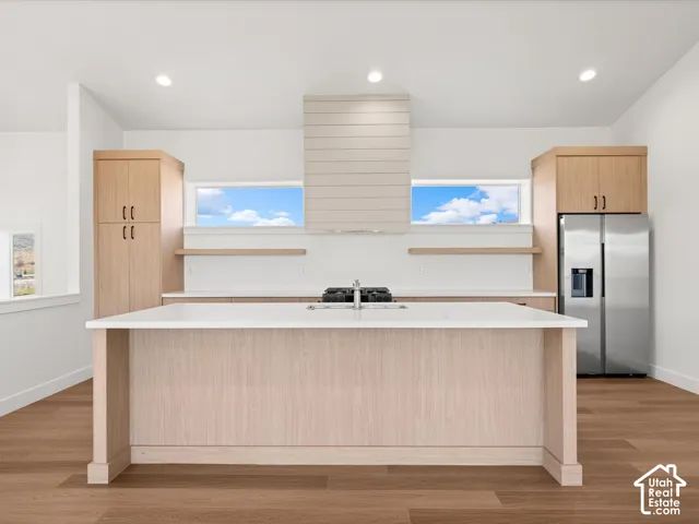 Kitchen featuring an expansive island with a white countertop, wood-finish flooring, light wood cabinetry, and a stainless steel refrigerator