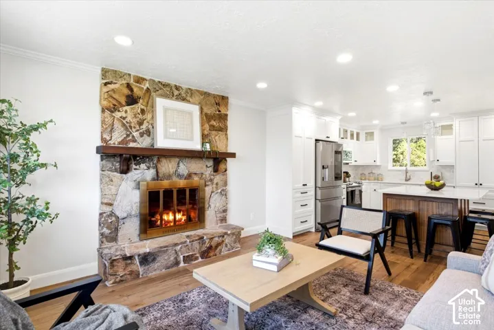 Stone-clad fireplace with wood mantle, wood-finish flooring, recessed lighting, and a kitchen featuring white cabinetry, stainless steel appliances, and an island with seating