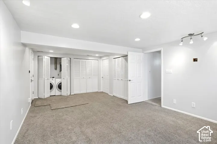 Recessed lighting, track lighting, light grey walls, grey carpet, and a laundry area with stacked appliances