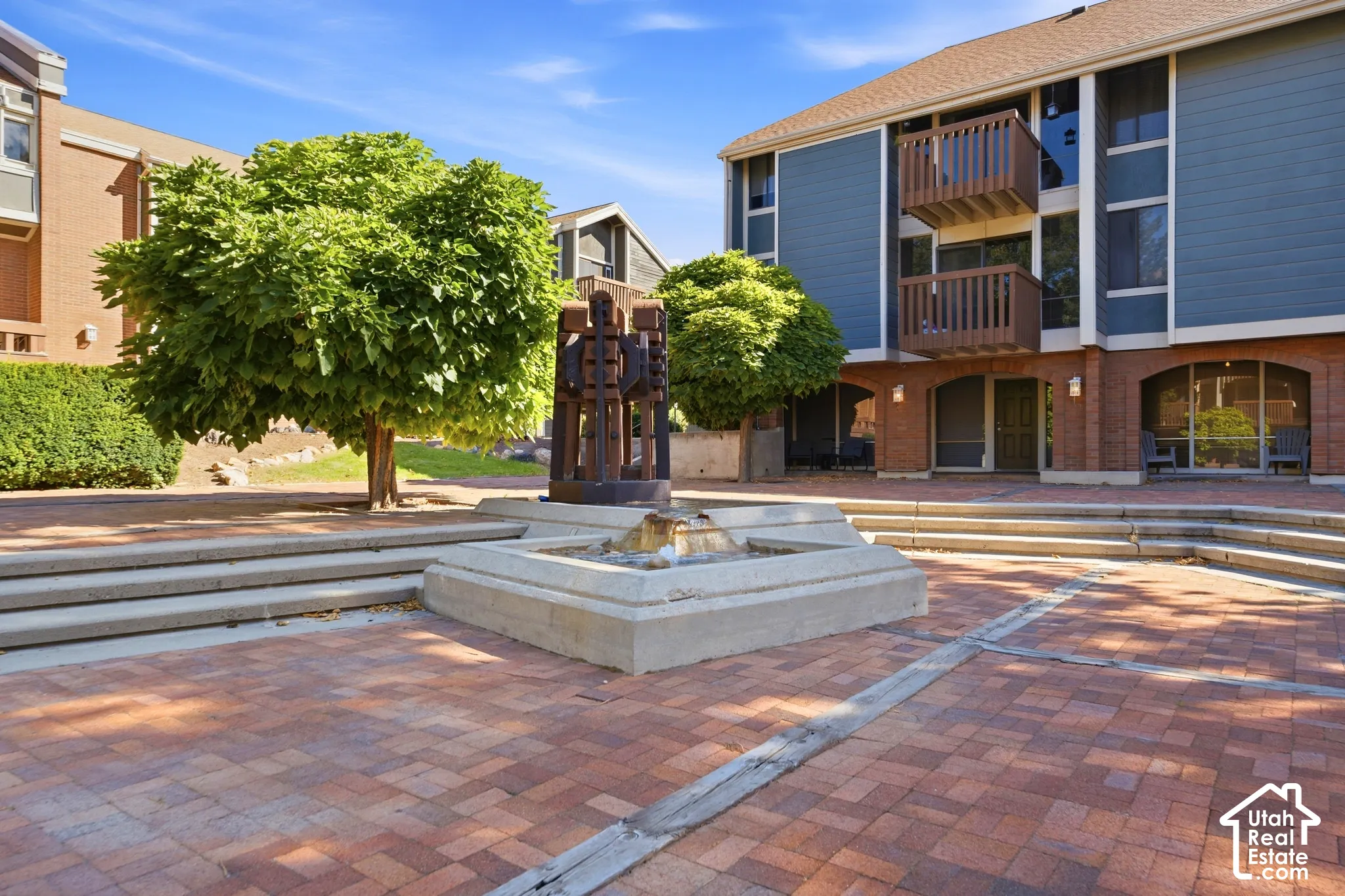 Brick paver courtyard with a central fountain, surrounded by mature trees and multi-level buildings featuring balconies