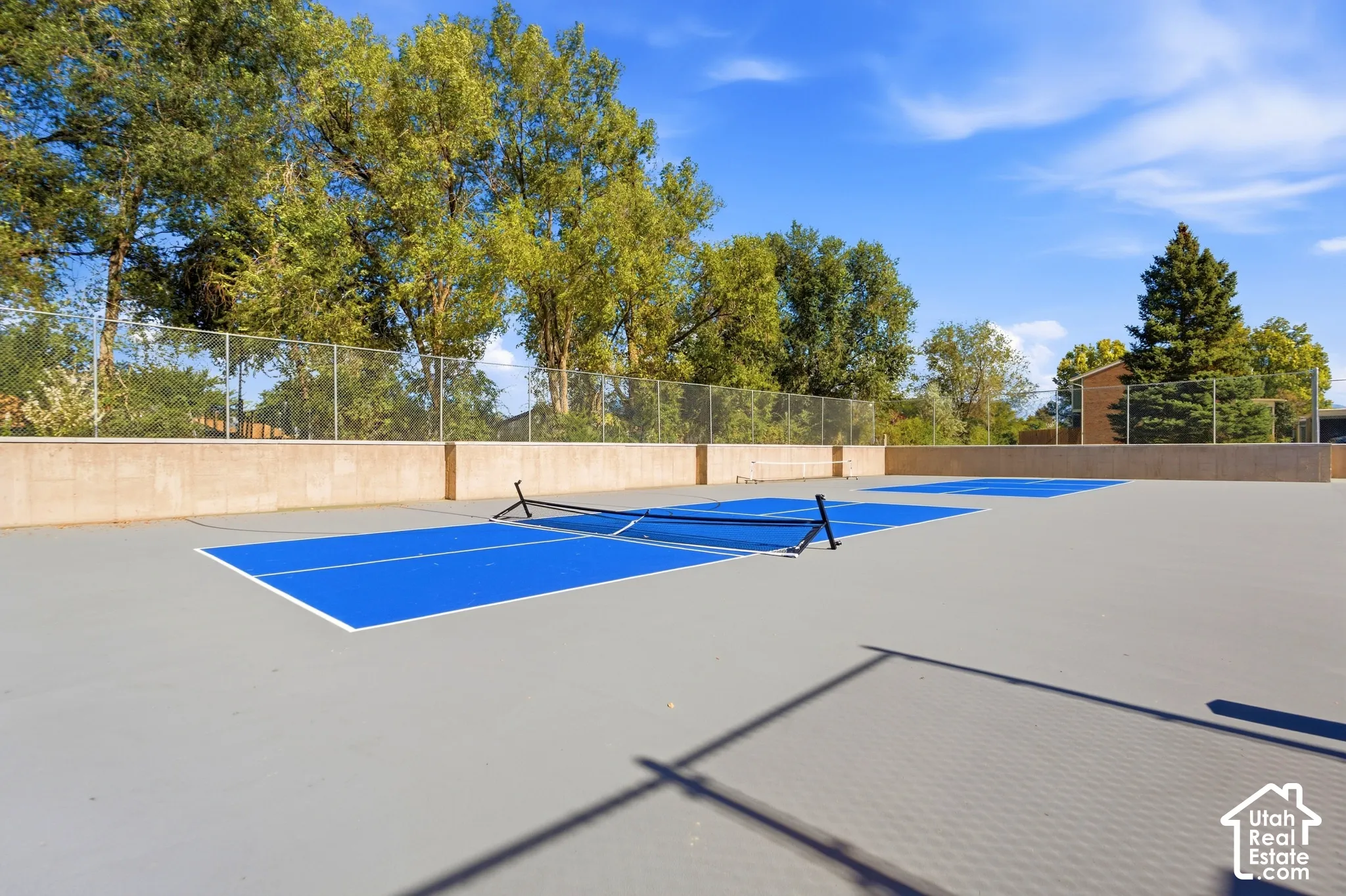 Two pickleball courts with blue playing surfaces and a gray surrounding area, enclosed by a perimeter fence and a low retaining wall