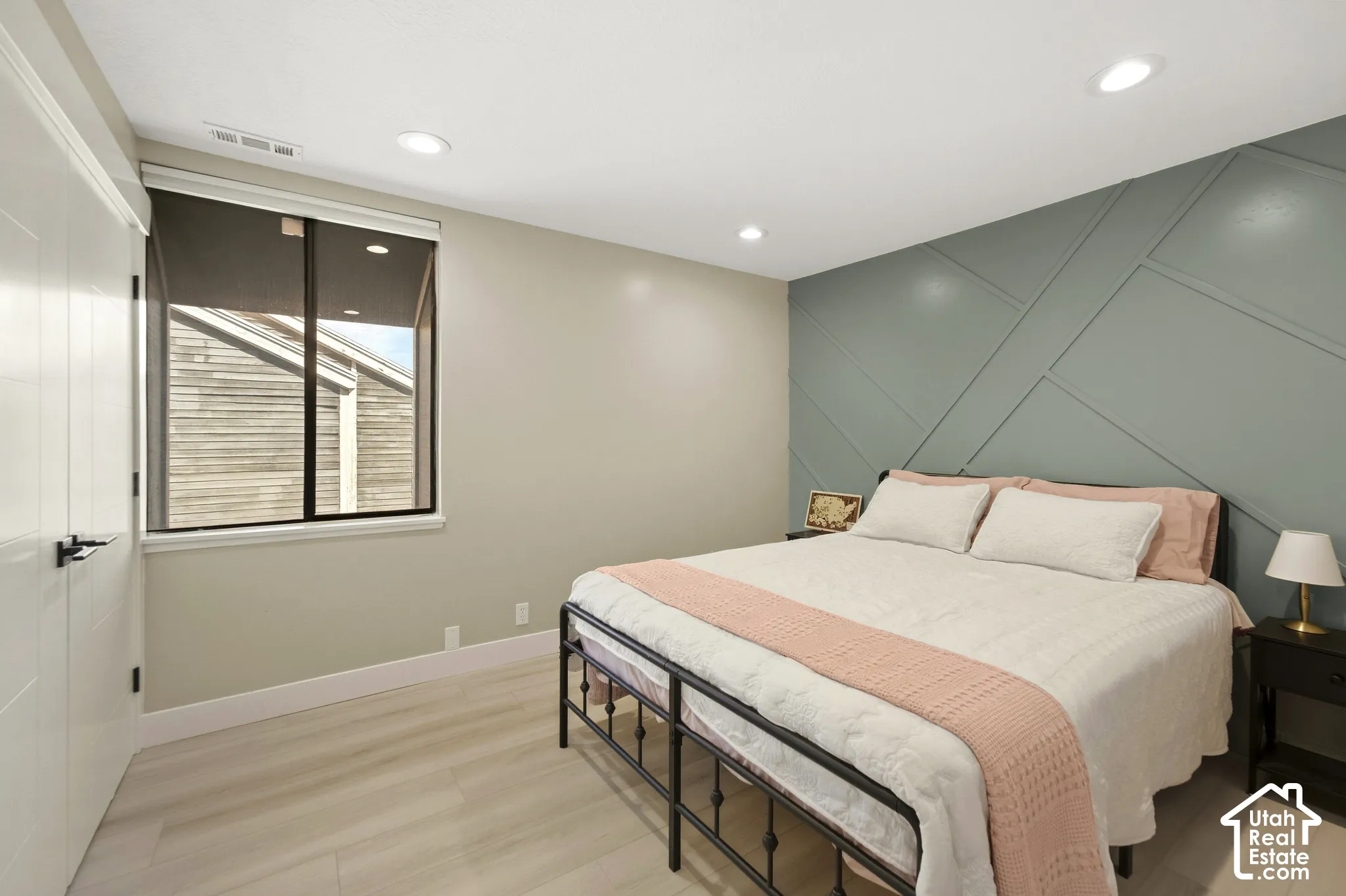 Bright bedroom with recessed ceiling lighting, large fixed window with blinds, light wood-finish flooring, and an accent wall featuring geometric paneling in soft sage green