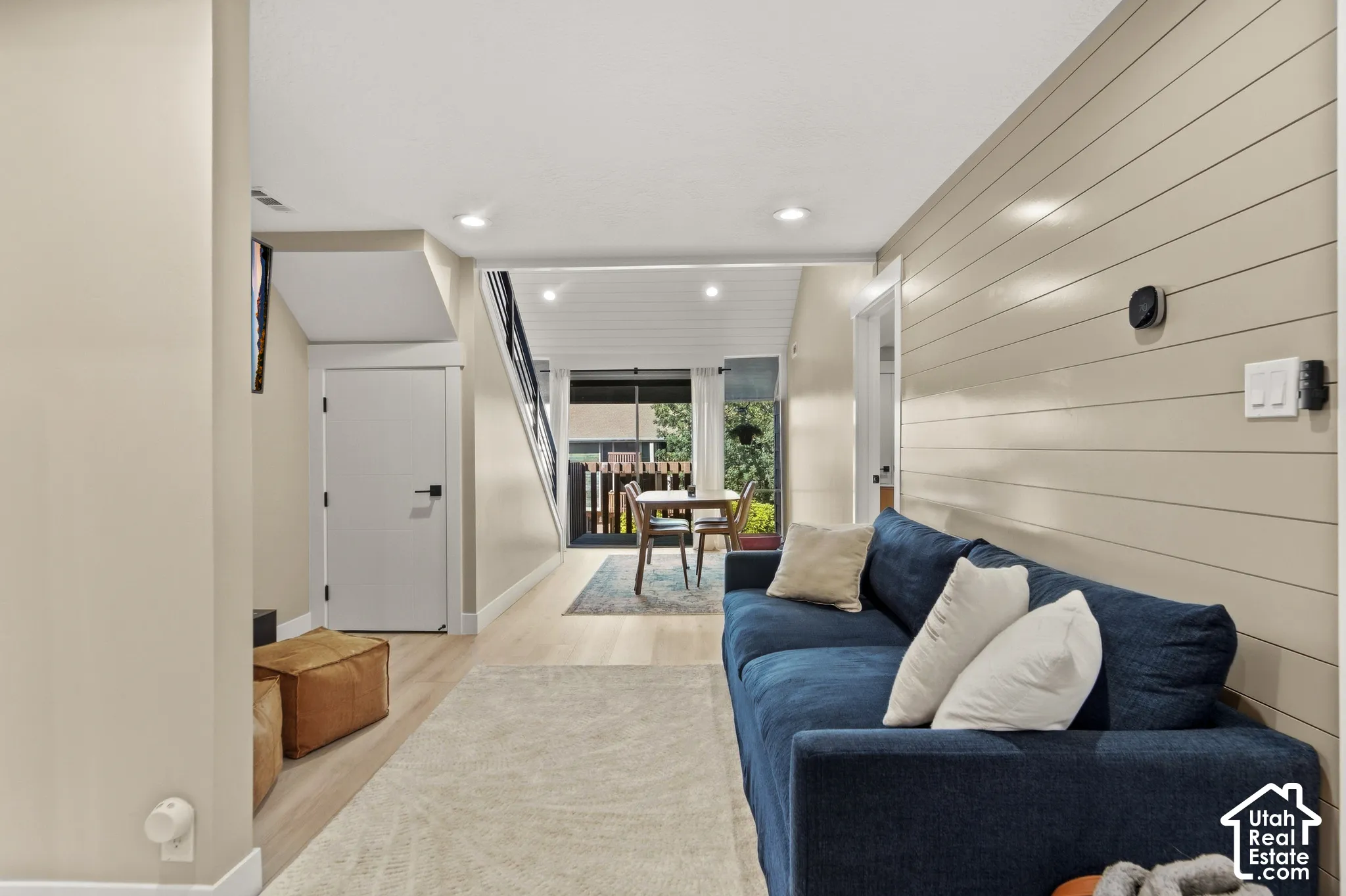 Recessed lighting illuminates the open living area featuring wood-finish flooring, a shiplap accent wall, and a staircase with dark railings