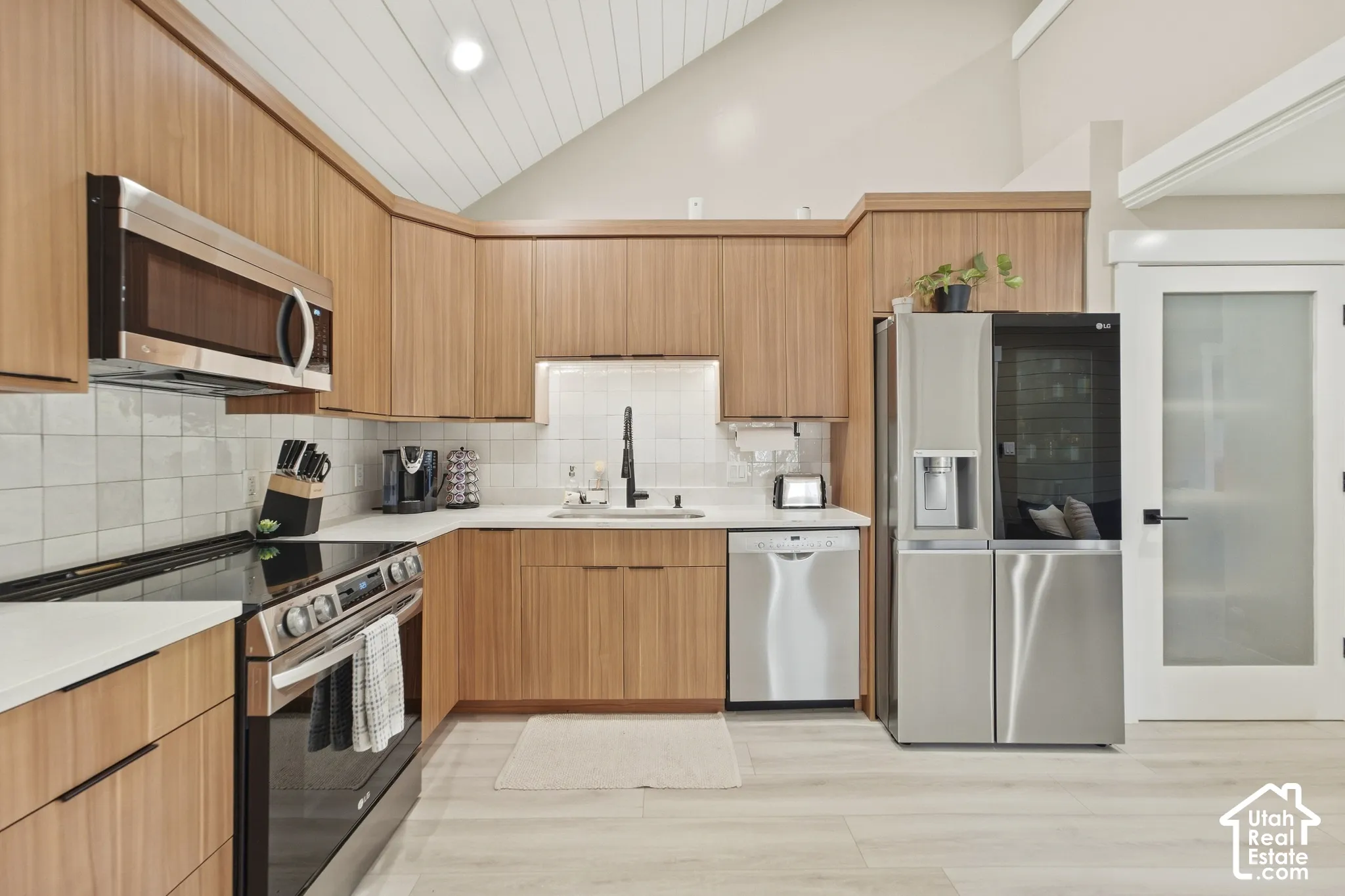 Kitchen featuring light wood-finish cabinetry, stainless steel appliances, a white tile backsplash, light-colored countertops, and wood-finish flooring