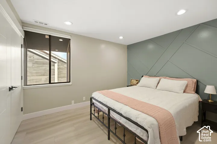 Bright bedroom with recessed ceiling lighting, large fixed window with blinds, light wood-finish flooring, and an accent wall featuring geometric paneling in soft sage green