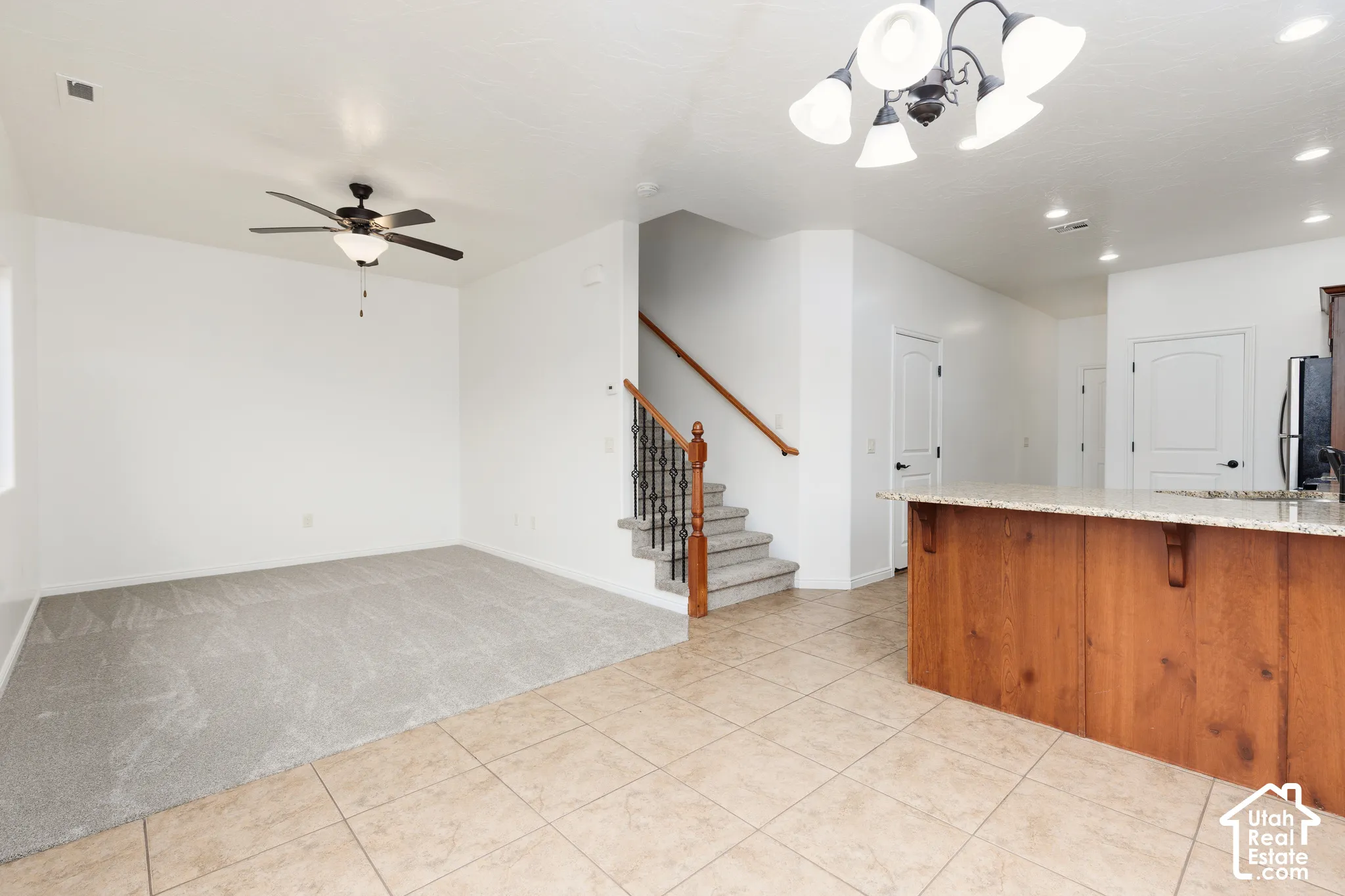 Ceramic tile flooring, carpeted living area, ceiling fan with integrated lighting, wood-finish kitchen island with stone countertop, and staircase with wrought iron balusters