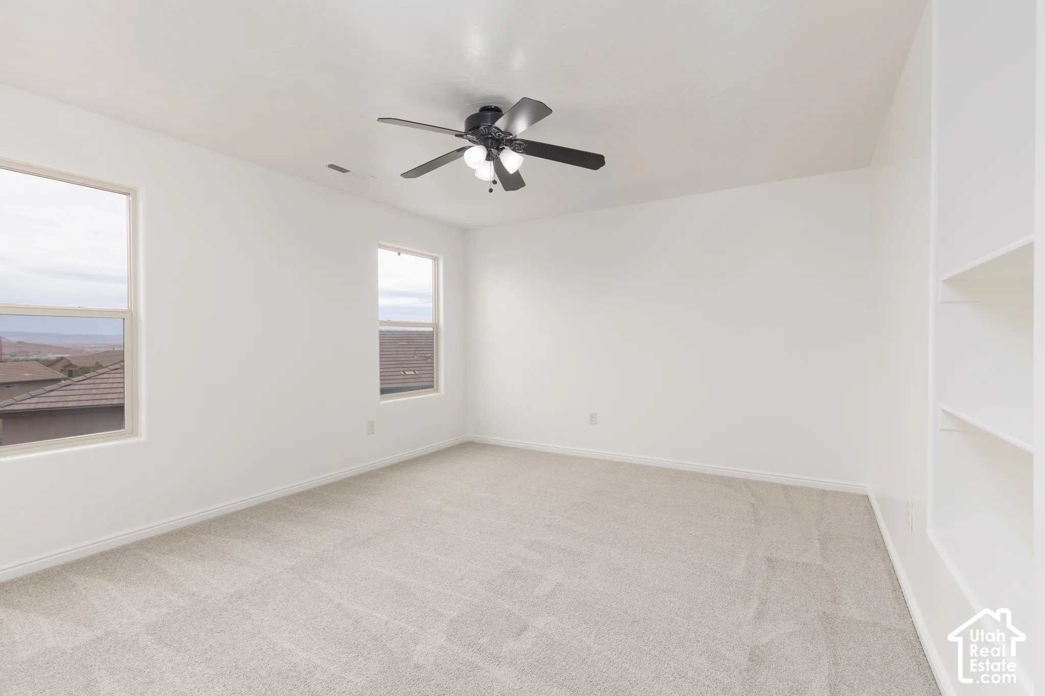 Neutral tone wall paint, light-colored carpeting, multiple windows, built-in shelving, and a ceiling fan with integrated lighting