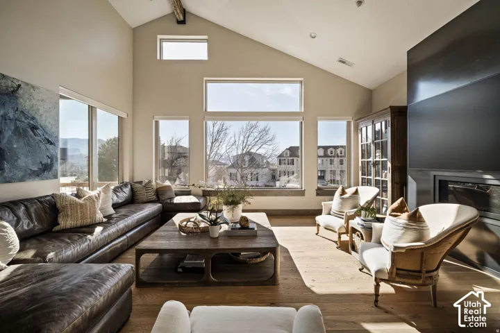 Vaulted ceiling with an exposed wood beam, wood-finish flooring, multiple large windows, and a built-in fireplace with a dark surround