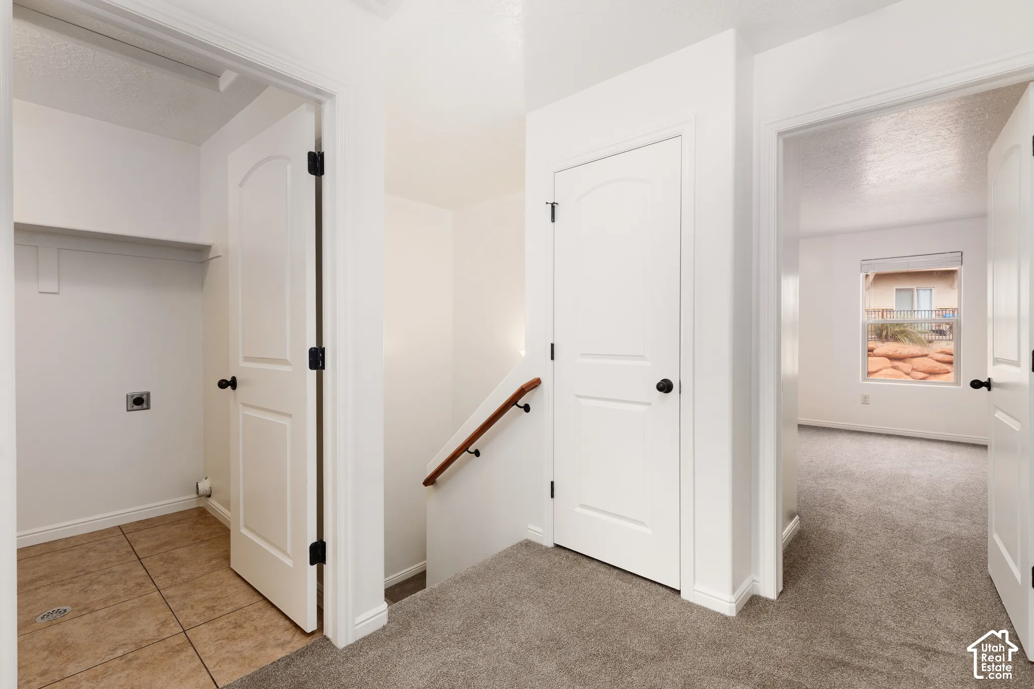 Laundry area with tiled flooring and storage shelf, hallway with carpeted flooring, staircase with wood handrail, and white interior doors with black hardware