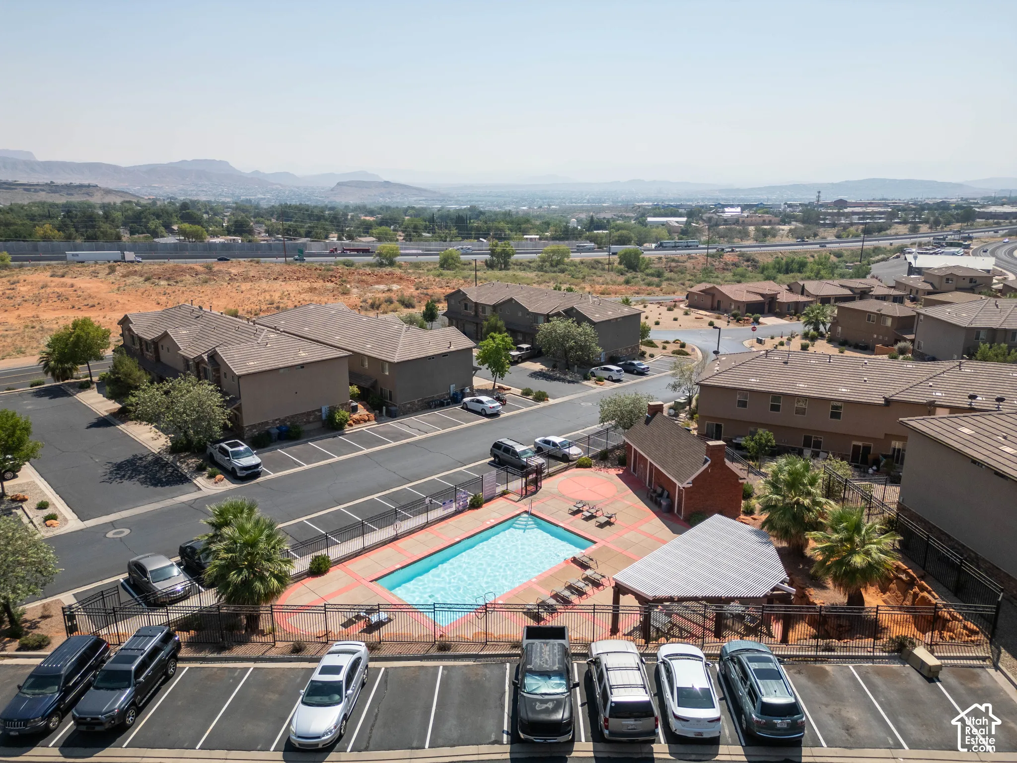 Community swimming pool with surrounding fence, adjacent covered seating area, and a large paved parking lot
