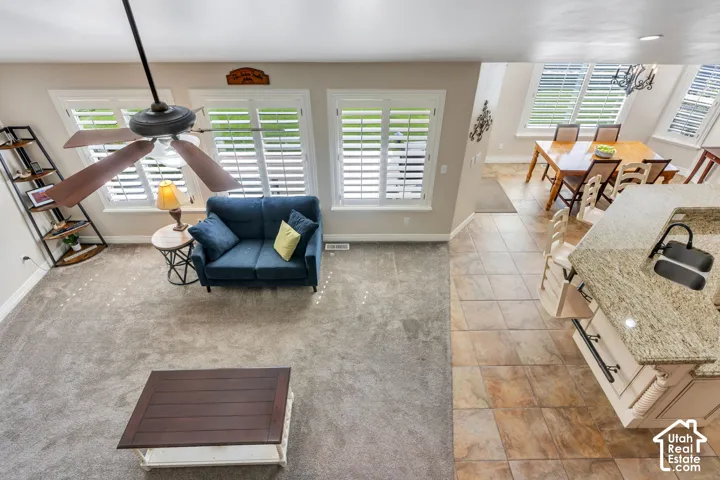 Open-concept living space with carpeted flooring, wood-finish ceiling fan, and multiple windows with plantation shutters