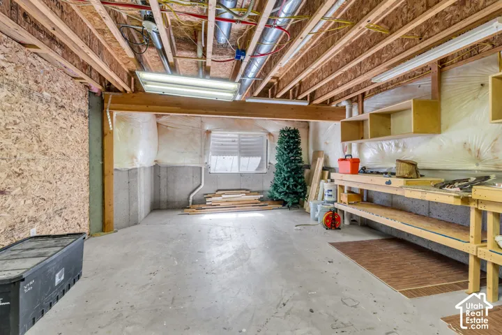 Unfinished workshop space in the basement. Concrete flooring, exposed ceiling joists, concrete foundation walls, built-in wooden shelving, and fluorescent lighting