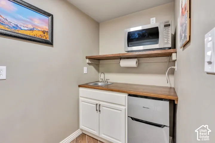 Theater room kitchenette featuring wood-finish countertops, white cabinetry, an under-counter refrigerator, a stainless steel sink with a chrome faucet, and an overhead wood-finish shelf