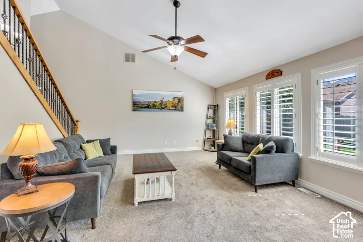 Vaulted ceiling with ceiling fan, three windows with white louvered shutters, carpeted flooring, and a staircase featuring a wood handrail and wrought iron balusters