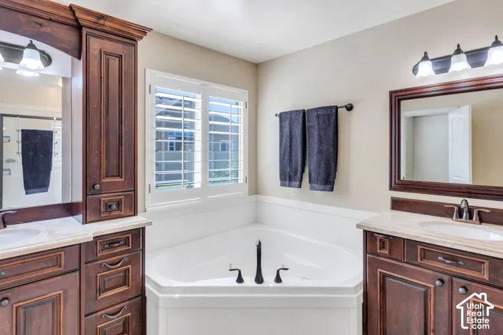 Corner soaking tub with dark bronze fixtures, dual wood-finish vanities with light stone countertops, white plantation shutters, and dark bronze light fixtures