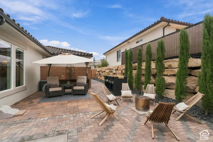 Paver patio featuring a built-in outdoor kitchen, fire pit, and tiered rock landscaping with privacy trees