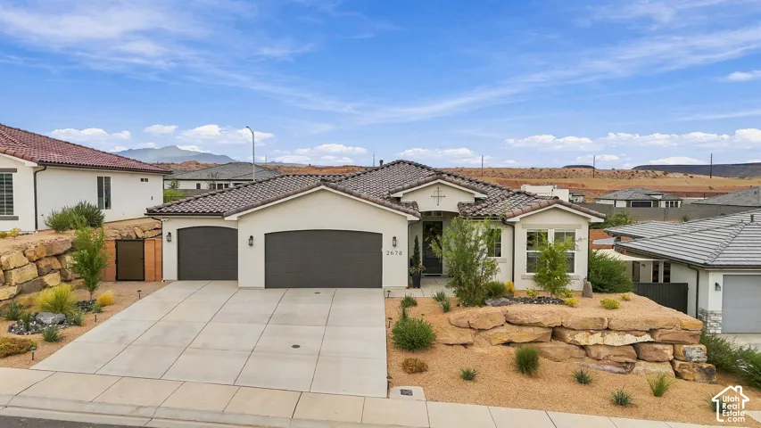 Stucco exterior with a tiled roof, two-car garage, concrete driveway, and desert-style landscaping featuring natural rock elements