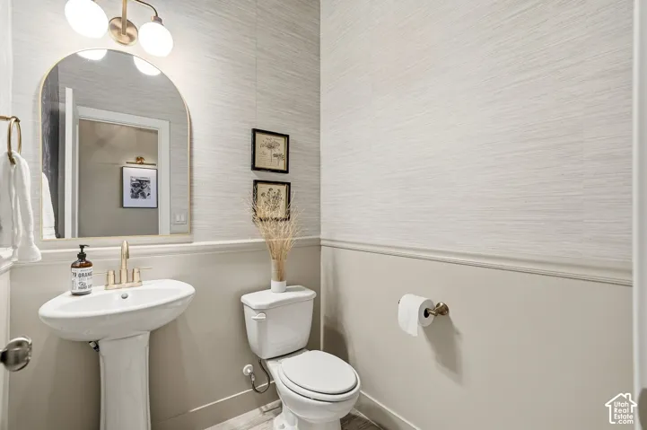 Pedestal sink with brass-finish faucet, arched mirror, brass-finish vanity light, and wainscoting with textured wall covering