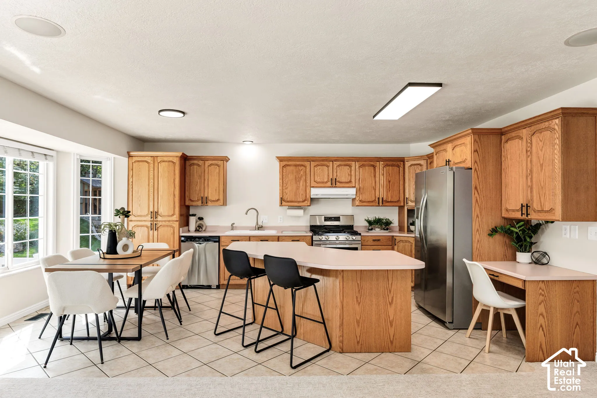 Open-concept kitchen with wood cabinetry, stainless steel appliances, a center island with counter seating, a dedicated desk area, and large bay windows