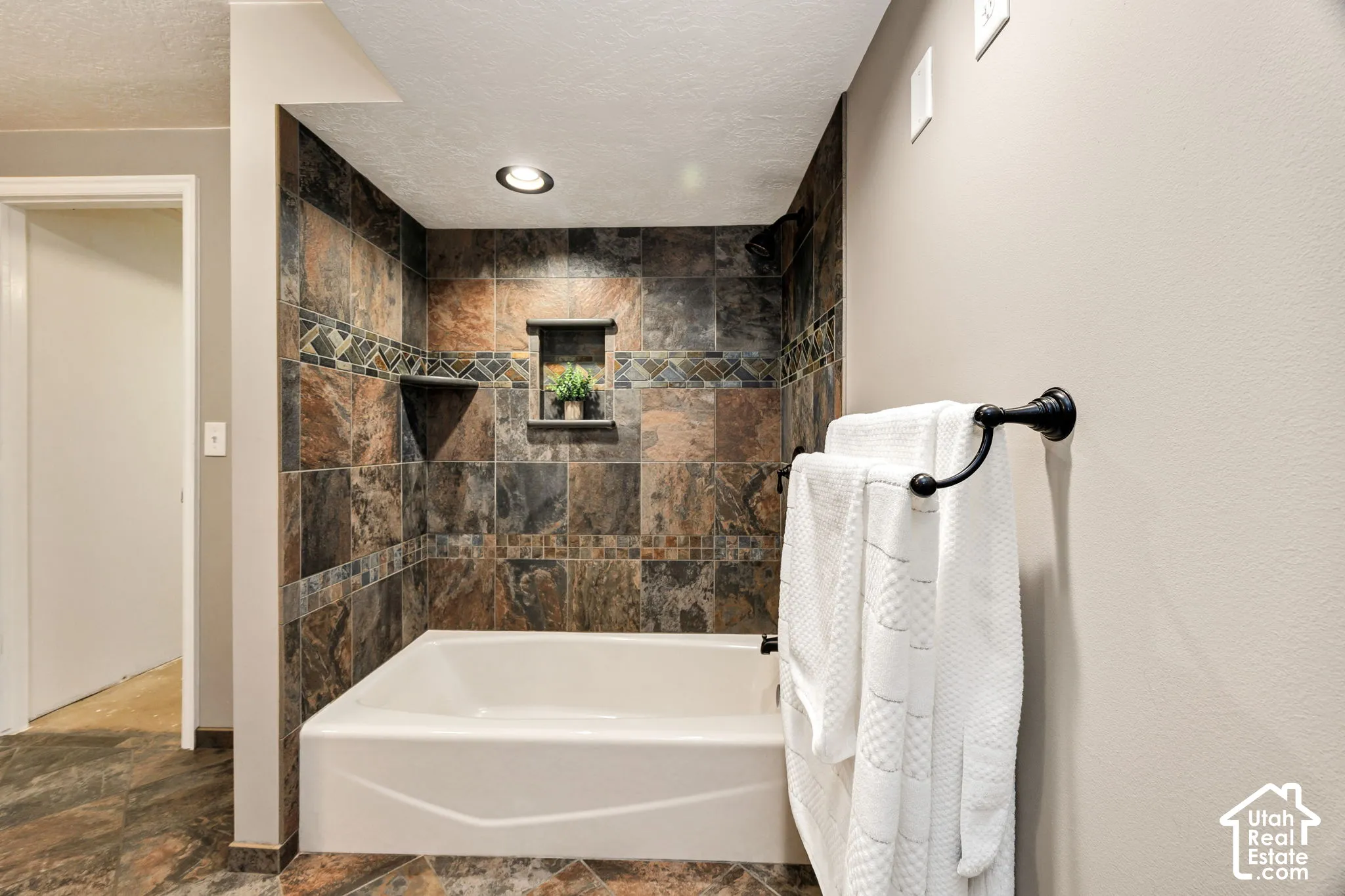 Bathtub with stone-finish tile surround, decorative tile accent band, recessed niche, and built-in shelving