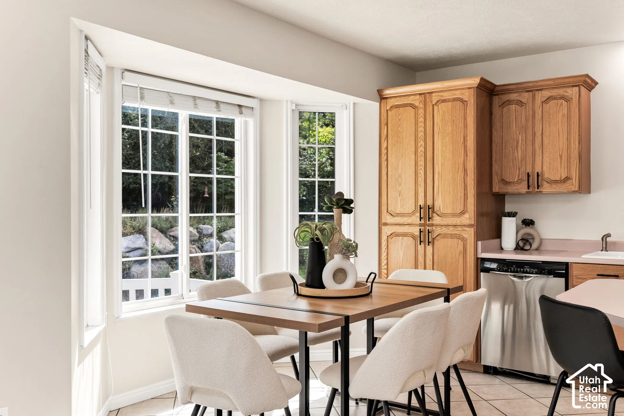 Bay window with grid-patterned windows, light wood-finish cabinetry, stainless steel dishwasher, and tiled flooring