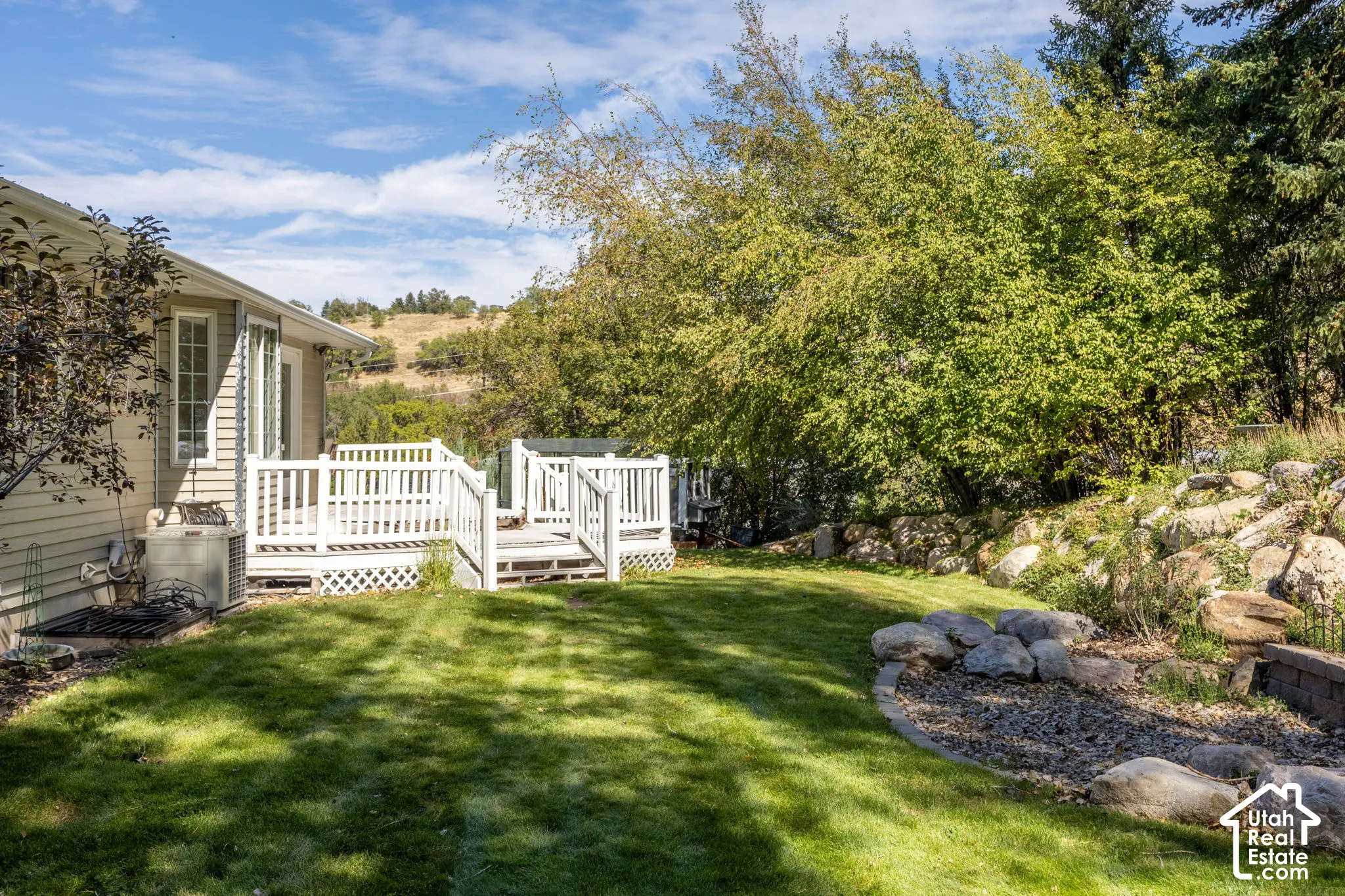 Expansive green lawn, white multi-level wood deck, rock landscaping, mature trees, and a distant hillside backdrop