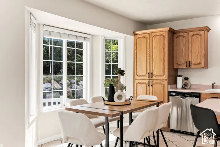 Bay window with grid-patterned windows, light wood-finish cabinetry, stainless steel dishwasher, and tiled flooring