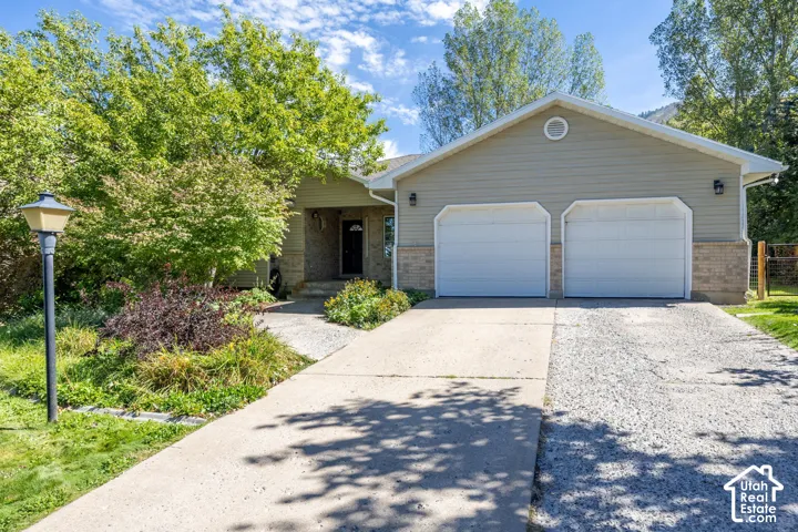 Light-colored siding exterior with a stone base, featuring a two-car garage, mature trees, and a concrete driveway