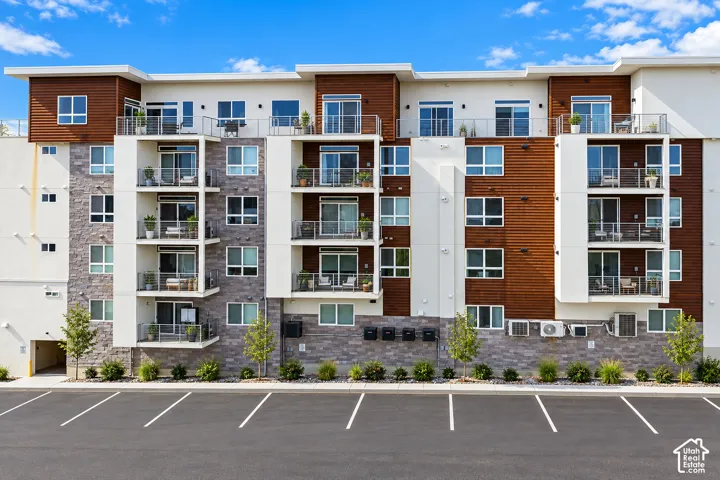 Modern building exterior with white and wood-finish siding, stone-look accents, multiple balconies with glass railings, and an adjacent asphalt parking lot