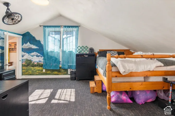 Sloped ceiling room featuring a window, textured carpeting, and a built-in ceiling fan