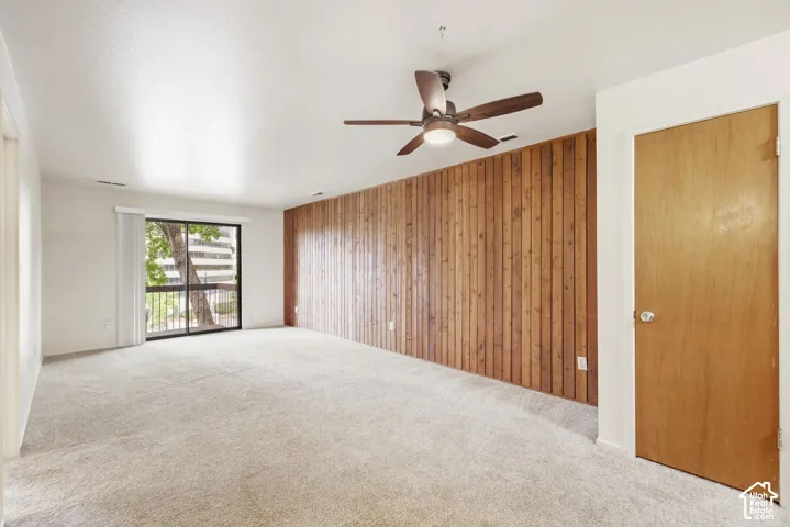 Carpeted flooring, wood panel accent wall, sliding glass door, ceiling fan with integrated lighting, and a solid wood door