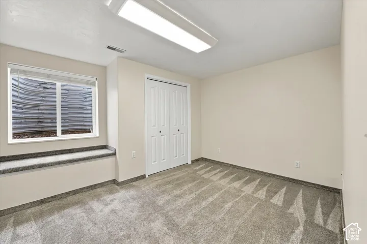 ADU Basement - Bright, light-filled bedroom featuring neutral carpeting, a large window with horizontal blinds, dual closets with louvered doors, and recessed ceiling lighting throughout