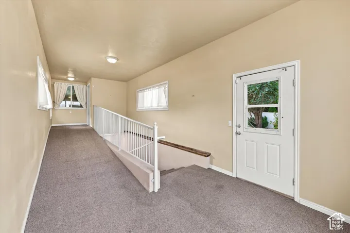 Bright upper-level hallway featuring neutral carpeting, white railing, recessed lighting, and direct access to exterior entry door with natural light from multiple windows