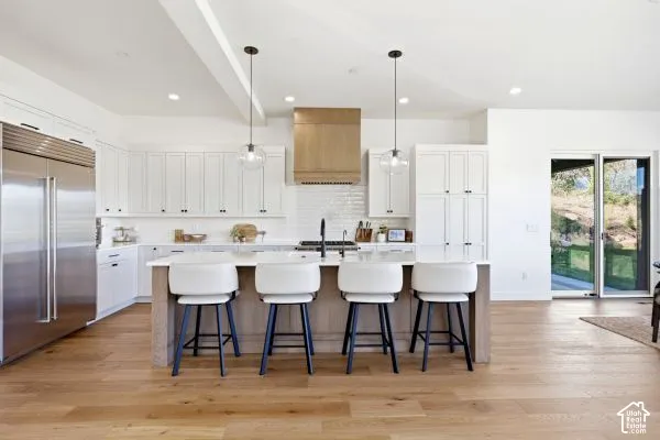 Open-concept kitchen featuring wood-finish flooring, white cabinetry, a large center island with seating, stainless steel appliances, and a light wood-finish range hood