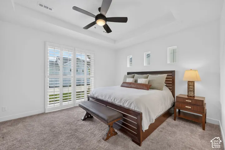 Tray ceiling with a ceiling fan, white plantation shutters, three high rectangular windows, and neutral carpeting