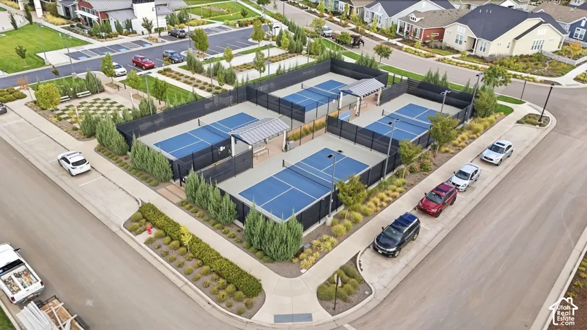 Four blue-surfaced courts with black fencing, two covered seating areas, surrounding landscaped grounds, and a paved pathway