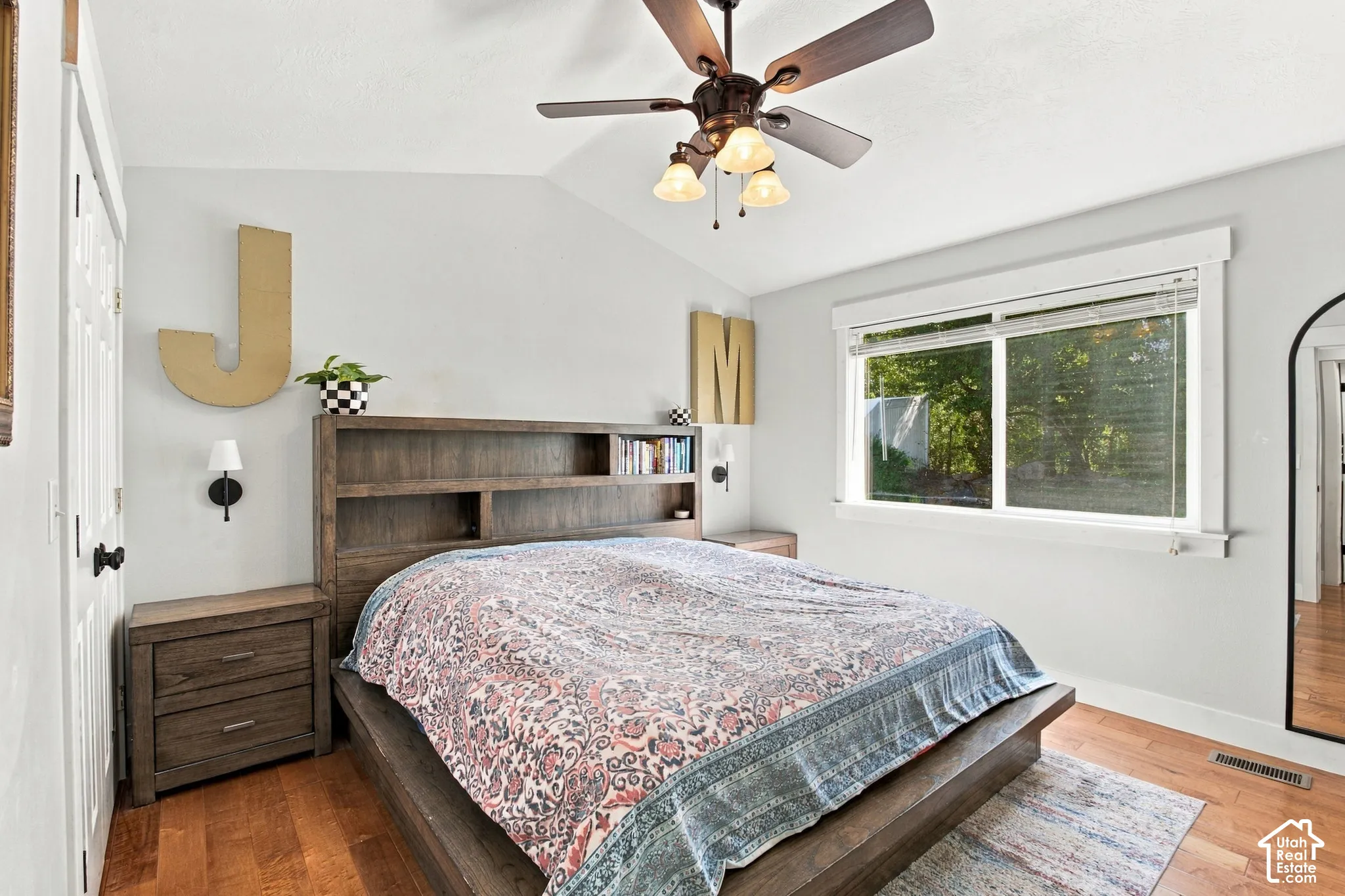 Primary Bedroom with sloped ceiling with a ceiling fan, wood-finish flooring, built-in headboard shelving, wall-mounted sconces, and a large window