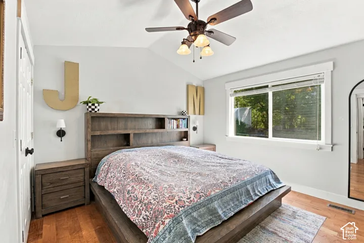 Primary Bedroom with sloped ceiling with a ceiling fan, wood-finish flooring, built-in headboard shelving, wall-mounted sconces, and a large window