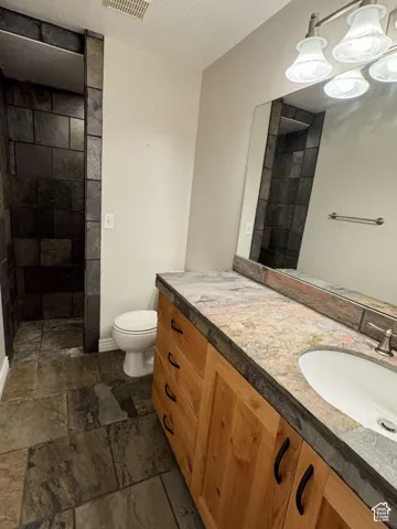Walk-in shower with natural stone tile, natural stone tile flooring, natural wood vanity with integrated sink, natural stone countertop, and vanity mirror with mounted lighting