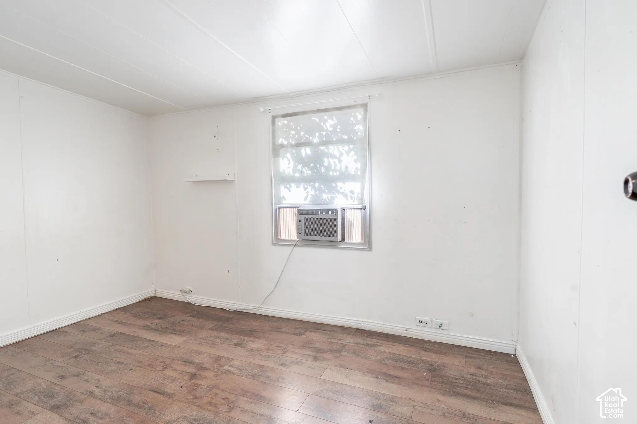 Wood-finish flooring with white panel walls, featuring a window-mounted air conditioning unit and a small wall-mounted shelf