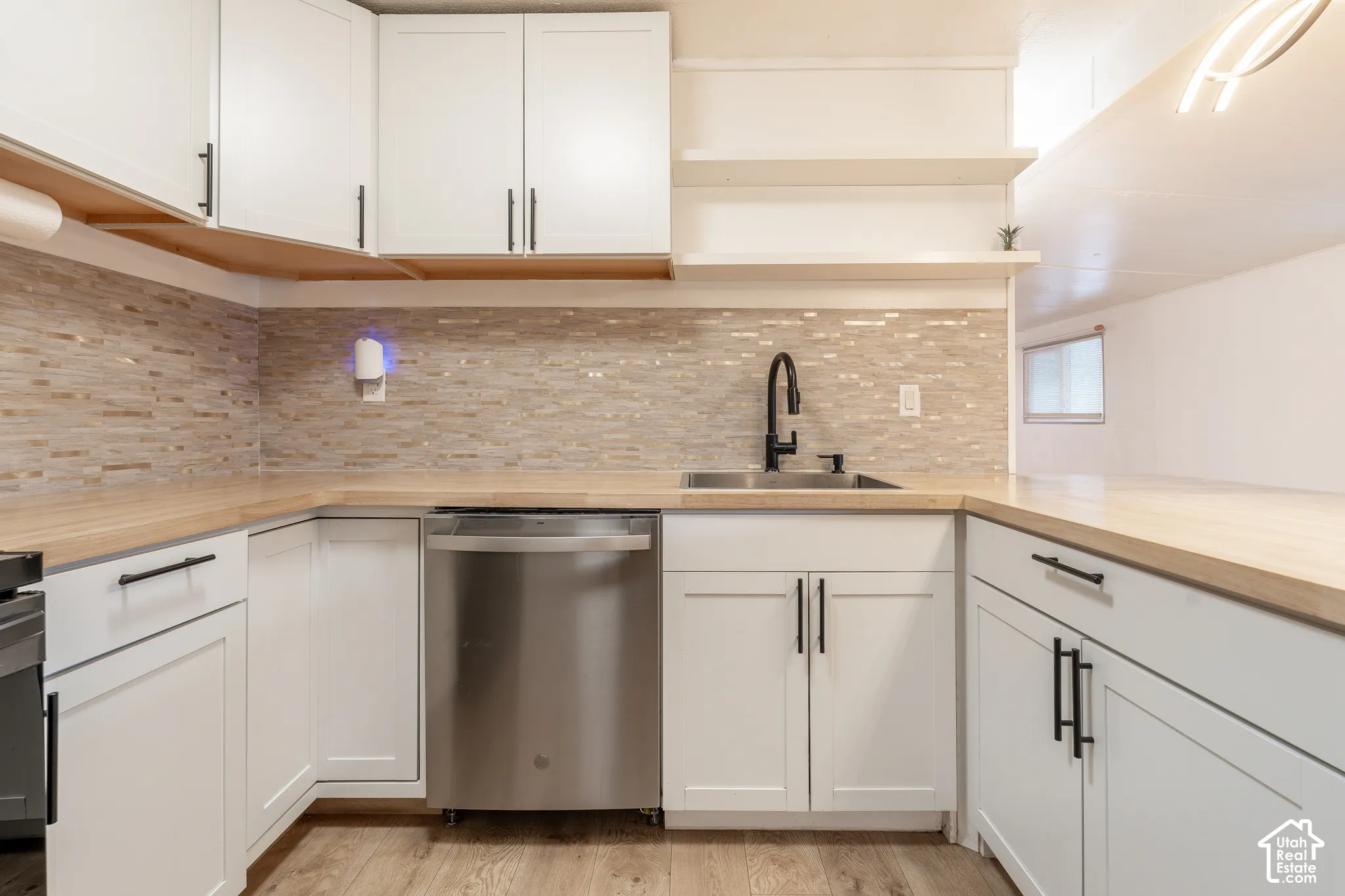 White cabinetry with matte black hardware, wood-finish countertops, stainless steel dishwasher, undermount sink with a matte black faucet, and a tiled backsplash